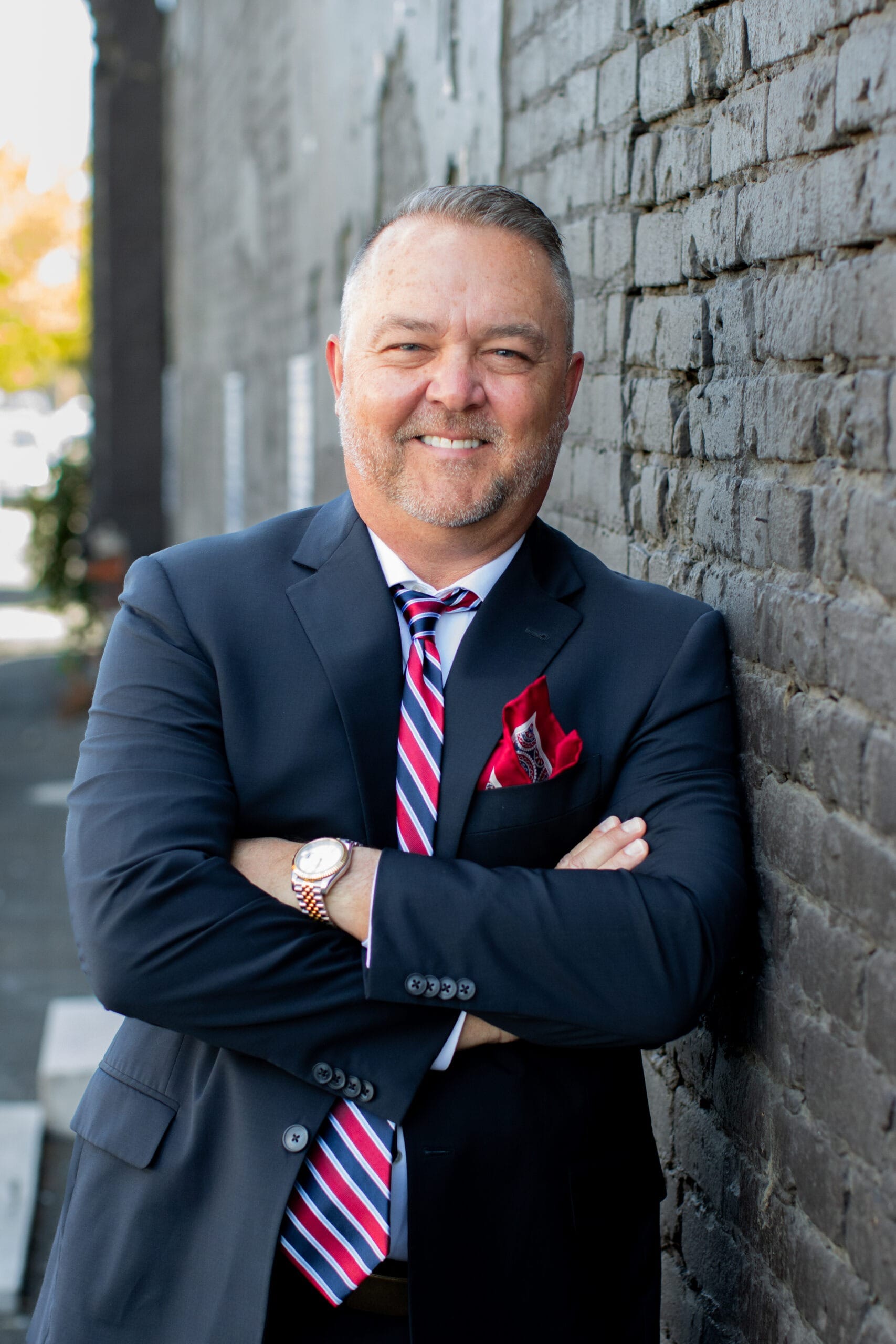 Confident man in a navy suit with a striped tie and red pocket square smiles while leaning against a gray brick wall, showcasing a professional demeanor.