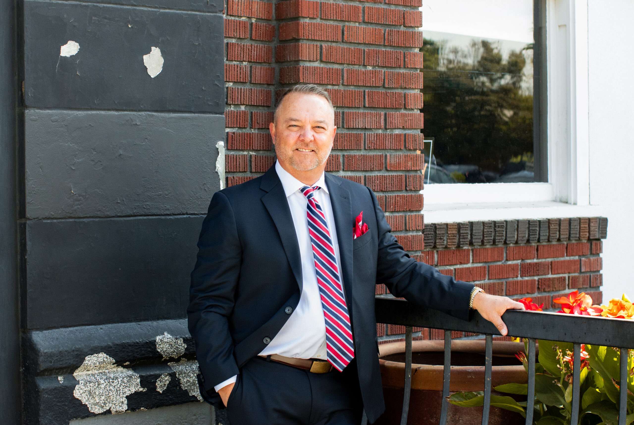 A well-dressed man in a dark suit with a striped tie and red pocket square leans against a brick wall, showcasing a friendly smile.