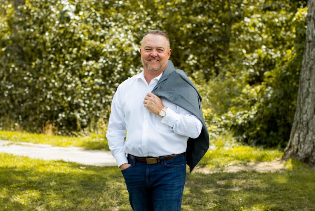 A smiling man casually dressed in a white shirt and jeans stands outdoors, holding a gray blazer. The background features lush greenery.